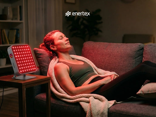 Una mujer relajada con los ojos cerrados en un sofá, recibiendo una sesión de terapia de luz roja de un panel de Envertex en un salón acogedor